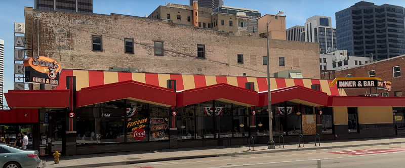 Street view of Sams 3 Diner & Bar with a retro-style red and yellow awning, a large brick building behind it, and a sign reading "Sams 3 Diner & Bar" along with a "DINER & BAR" sign on the right side.