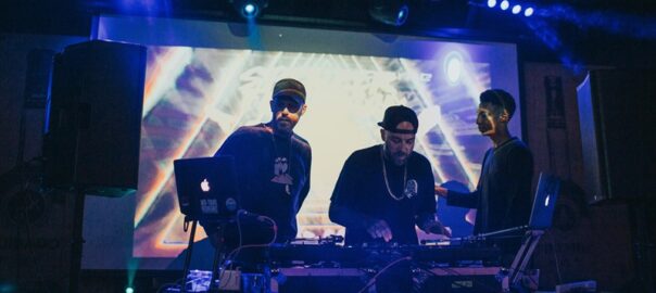 Three DJs performing live at a vibrant Denver music event with colorful lights and visuals in the background