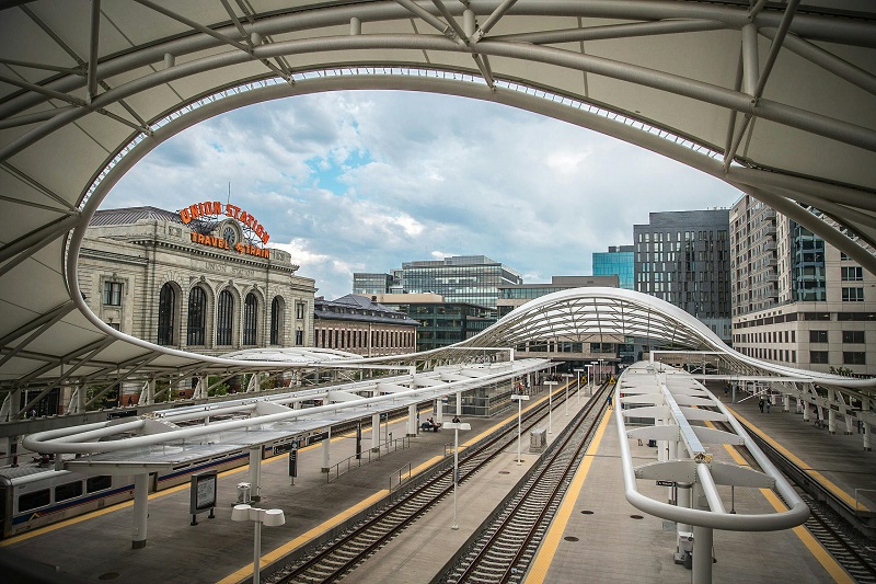 Modern railway platform with curved canopy roof at Denver Union Station, surrounded by historic and modern buildings under a cloudy sky.