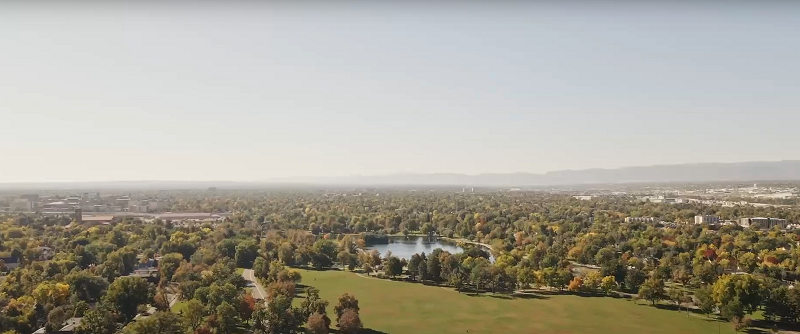 Aerial view of a large city park with green trees, a lake, and open grassy areas surrounded by urban buildings and distant mountains under a clear sky.