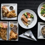 A variety of Denver’s signature dishes, including steak, fish, salad, tacos, and fries, beautifully plated on a table with wine bottles.