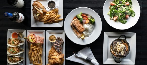 A variety of Denver’s signature dishes, including steak, fish, salad, tacos, and fries, beautifully plated on a table with wine bottles.